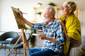 Older man painting while his wife stands behind him watching